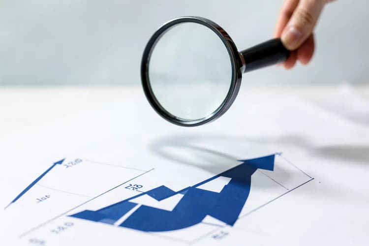Uma mão segurando uma lupa sobre um gráfico financeiro com setas apontando para cima.